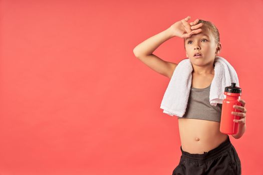 Adorable female child with towel on her shoulders holding bottle of water while standing against red background