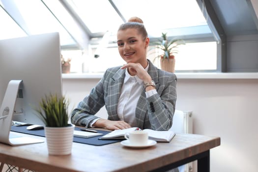 Portrait of a cheerful young businesswoman sitting at the table in office and looking at camera