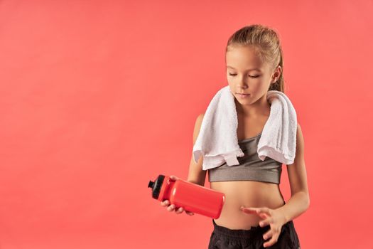 Adorable female child with towel on her shoulders looking at sports drink with serious expression while standing against red background