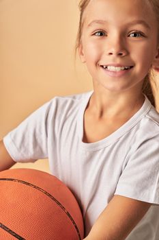 Close up of cute female child with basketball ball standing against light orange background