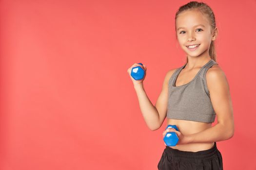Cute joyful girl in sportswear holding dumbbells and smiling while standing against red background