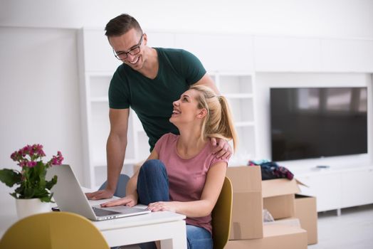 Young couple moving in a new home. Man and woman at the table using notebook laptop computer and plans with boxes around them