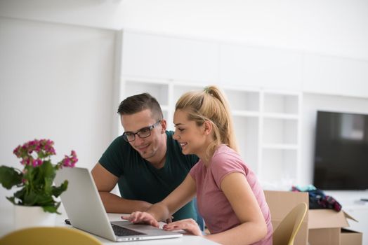 Young couple moving in a new home. Man and woman at the table using notebook laptop computer and plans with boxes around them