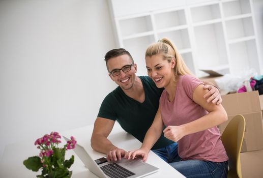 Young couple moving in a new home. Man and woman at the table using notebook laptop computer and plans with boxes around them