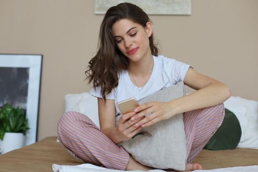 Beautiful young smiling brunette woman lying in white bed and using a phone in her bedroom.