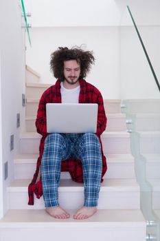 young freelancer in bathrobe working from home using laptop computer while sitting on stairs