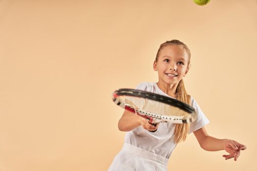 Adorable joyful girl tennis player hitting the ball with racket. Isolated on light orange background