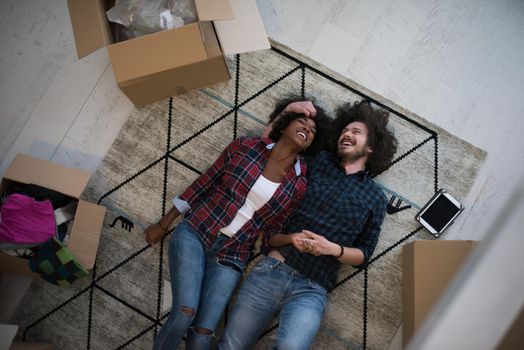 Top view of attractive young multiethnic couple moving, holding hands, looking at camera and smiling while lying among cardboard boxes