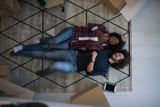 Top view of attractive young multiethnic couple moving, holding hands, looking at camera and smiling while lying among cardboard boxes