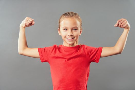 Adorable female child in red shirt showing her arm muscles and smiling while standing against gray background