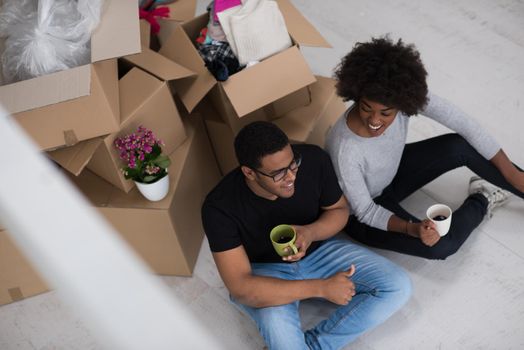 Relaxing in new house. Cheerful young African American couple sitting on the floor and drinking coffee while cardboard boxes laying all around them