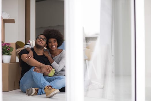 Relaxing in new house. Cheerful young African American couple sitting on the floor and drinking coffee while cardboard boxes laying all around them