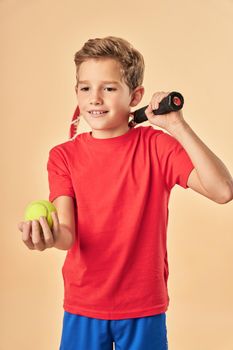 Cute male child tennis player holding yellow ball and smiling while standing against light orange background