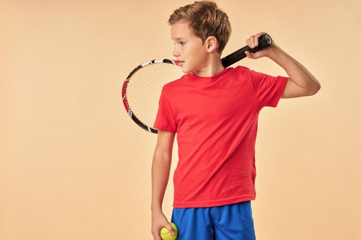 Adorable male child in red shirt holding tennis ball and racket while looking away with serious expression