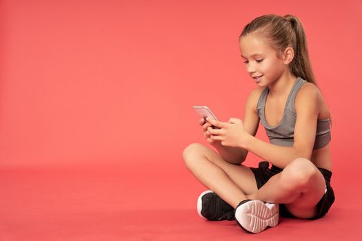 Cute female child in sportswear texting message on mobile phone and smiling