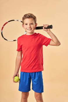 Adorable male child tennis player holding racket and smiling while standing against light orange background