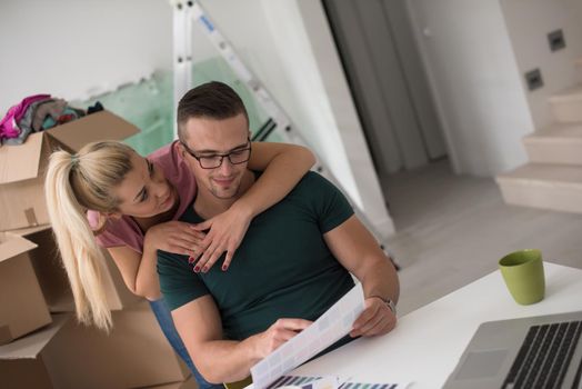 Young couple moving in a new home. Man and woman at the table using notebook laptop computer and plans with boxes around them