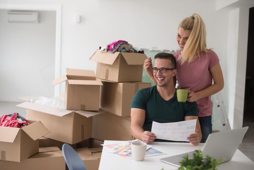 Young couple moving in a new home. Man and woman at the table using notebook laptop computer and plans with boxes around them