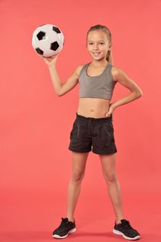 Adorable sporty girl in crop top and shorts looking at camera and smiling while holding football ball