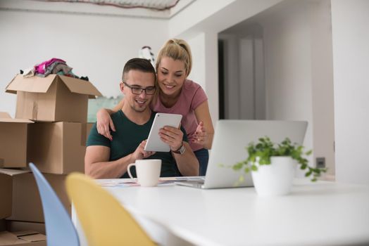 Young couple moving in a new home. Man and woman at the table using notebook laptop computer and plans with boxes around them