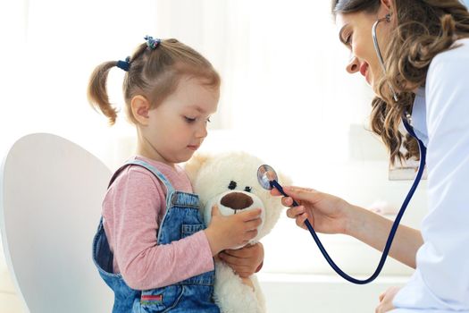 Little girl at the doctor for a checkup. Doctor playfully checking the heart beat of a teddy bear