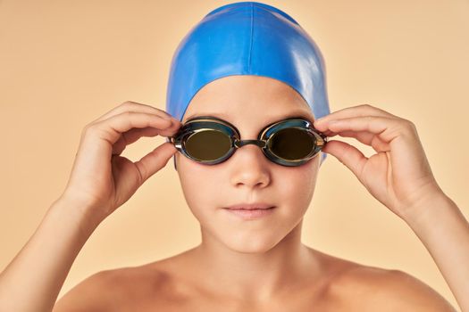 Close up of adorable boy swimmer in blue swim cap putting on goggles before swimming in pool