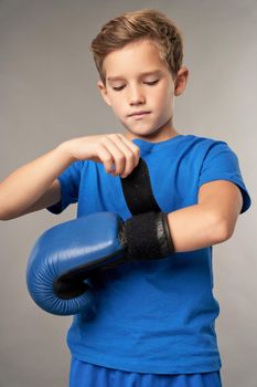 Serious cute boy preparing hands for boxing training while standing against gray background