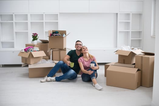 Relaxing in new house. Cheerful young couple sitting on the floor and drinking coffee while cardboard boxes laying all around them