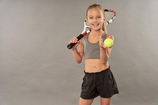 Adorable smiling girl in sportswear holding tennis racket and ball while standing against gray background