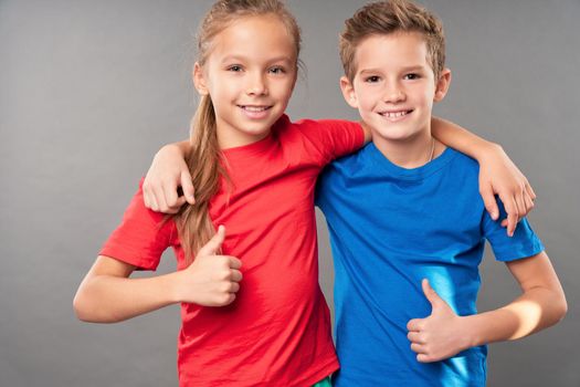 Adorable smiling kids showing approval gesture and hugging while standing against gray background
