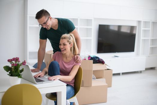 Young couple moving in a new home. Man and woman at the table using notebook laptop computer and plans with boxes around them