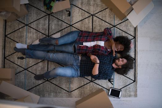 Top view of attractive young multiethnic couple moving, holding hands, looking at camera and smiling while lying among cardboard boxes