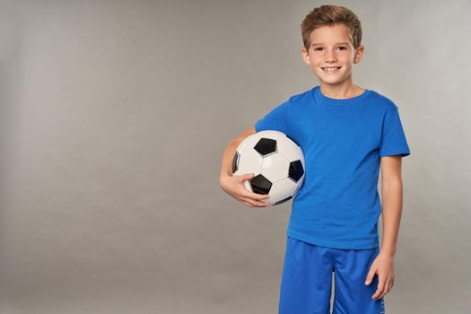 Joyful male child in blue shirt looking at camera and smiling while holding football ball
