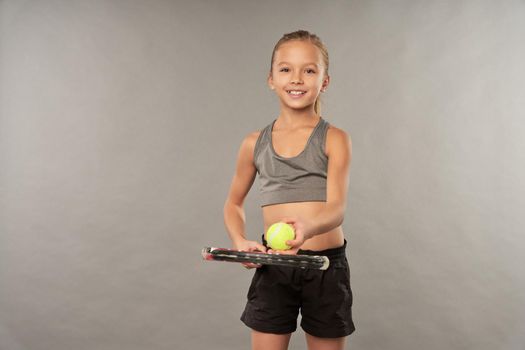 Cute smiling child holding tennis racket and ball while standing against gray background