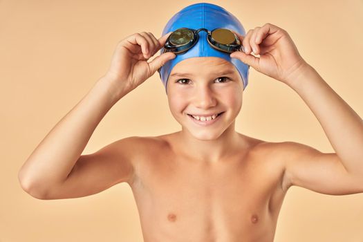 Close up of cheerful male child in swim cap looking at camera and smiling while adjusting swim goggles