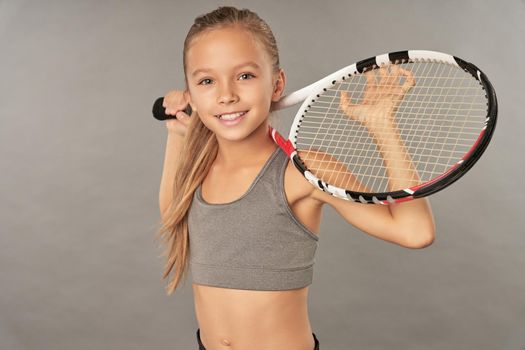 Cheerful female child tennis player looking at camera and smiling while holding racket behind her back