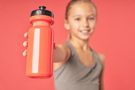 Close up of adorable female child with sports drink in her hand looking at camera and smiling while standing against red background