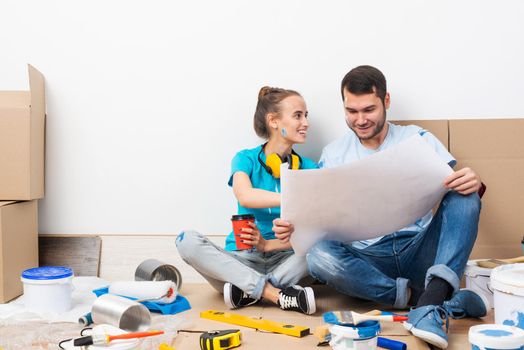 Young man and woman together planning their home renovation. Cardboard boxes, painting tools and materials on floor. House remodeling and interior renovation. People looking at blueprint at home.