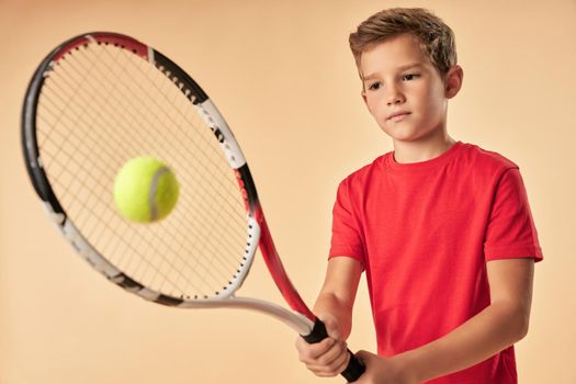 Cute male child tennis player hitting the ball with racket while standing against light orange background