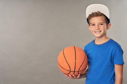 Joyful male child in cap holding basketball ball and smiling while standing against gray background