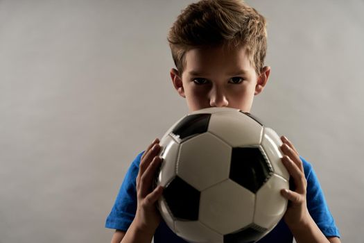 Cute boy holding football ball and looking at camera while standing against gray background