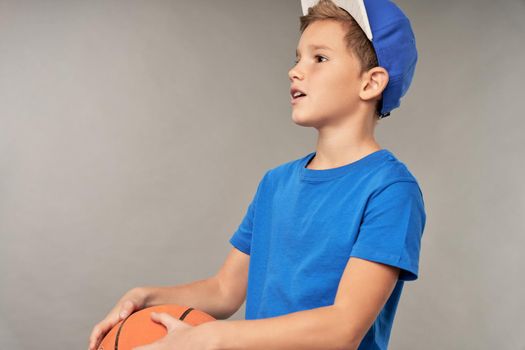 Adorable male child wearing blue shirt and cap while holding basketball ball and looking away