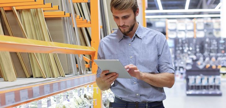 Young man shopping or working in a hardware warehouse standing checking supplies on his tablet