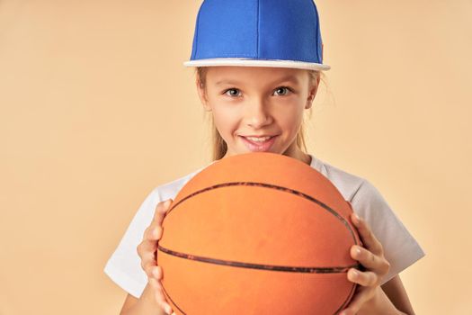 Cheerful female child basketball player holding game ball and smiling while standing against light orange background