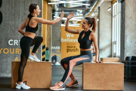 Great workout. Two young athletic fitness girls in sportswear giving high five each other while sitting on crossfit jump boxes at gym, exercising together. Sport, training and healthy lifestyle