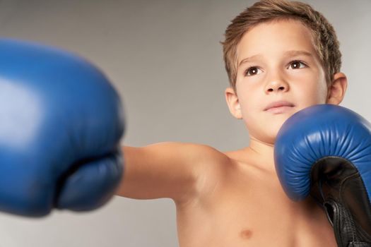 Cute male child in boxing gloves punching air and looking away while standing against gray background