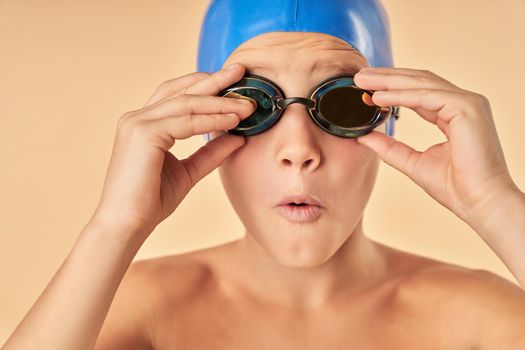 Close up of cute boy swimmer wearing swimming cap and glasses while standing against light orange background