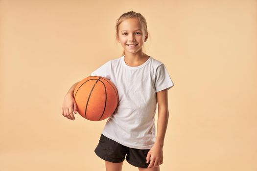 Adorable female child holding basketball ball and smiling while standing against light orange background