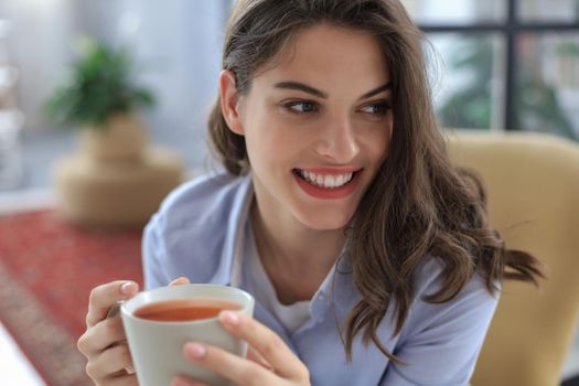 Smiling young female sitting in the armchair in the living room, holding a cup of coffee