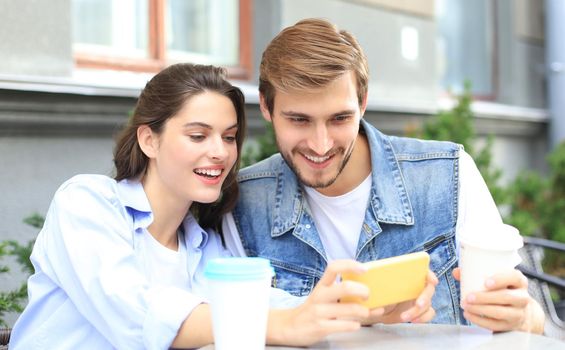 Attractive young couple in love drinking coffee while sitting at the cafe table outdoors, using mobile phone